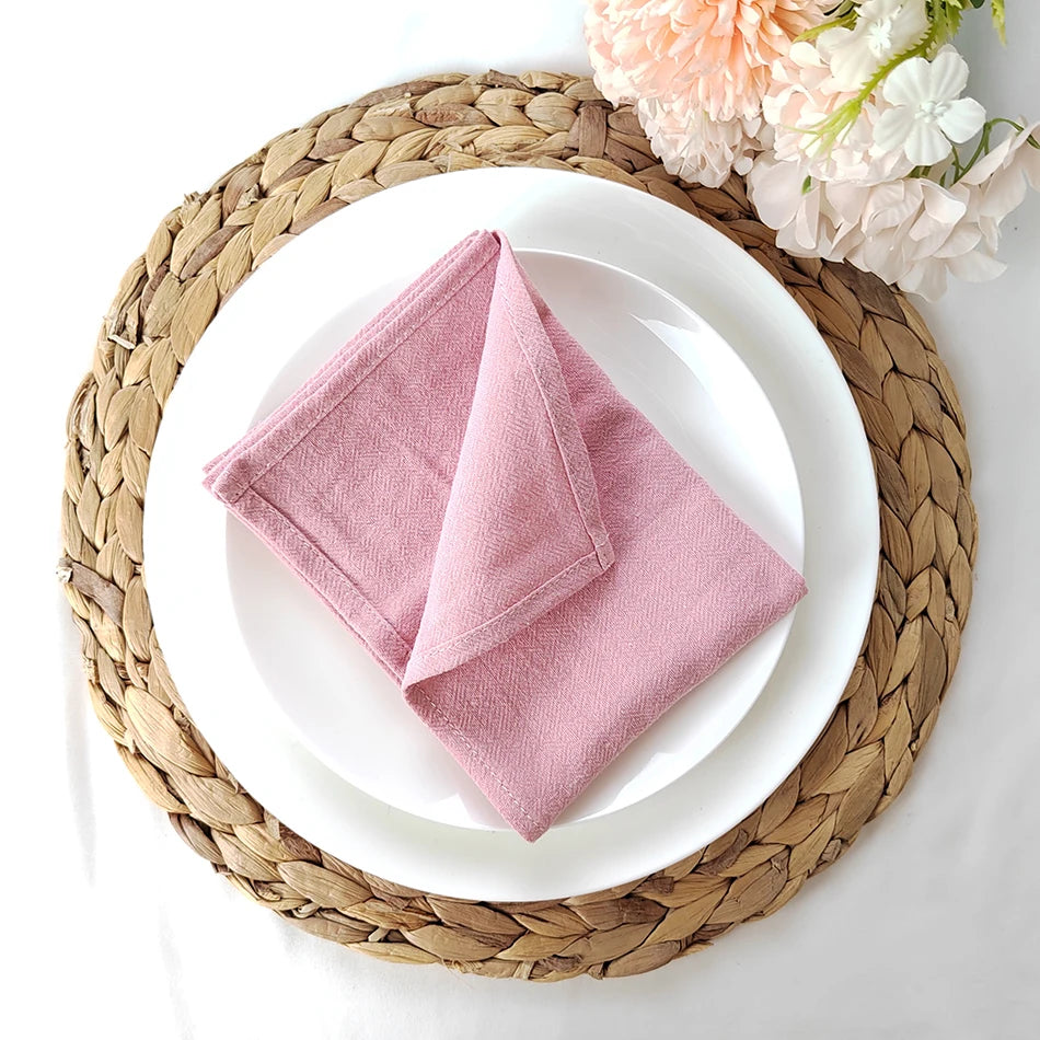 Une belle serviette en lin rose pastel posée sur une assiette blanche entourée d'un napperon en osier tressé, avec des fleurs de dahlia rose pâle à côté.