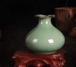 Un vase en céramique de couleur vert pâle posé sur un support en bois rougeâtre, créant un contraste harmonieux.