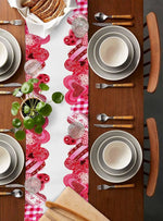 Cette image montre une table dressée avec des assiettes, des couverts, un chemin de table rose motif coeurs et une jolie plante verte, évoquant un moment de repas convivial et chaleureux.
