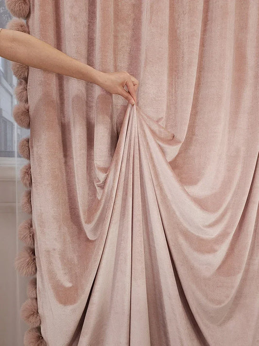 Main soulevant un rideau rose poudré en velours fluide, mettant en valeur son tombé souple et sa texture soyeuse.