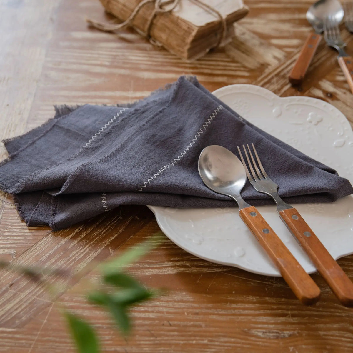 Sur une table en bois, une assiette blanche avec une fourchette et une cuillère en métal à manches en bois, entourée d'un linge de table en coton gris.