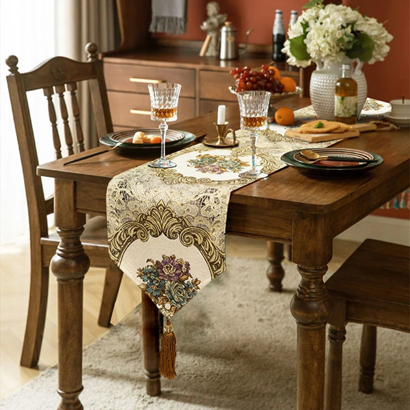 Cette image montre une table de salle à manger en bois, joliment décorée avec un chemin de table ornementé, des fleurs dans un vase, des verres à vin et de la nourriture sur la table.