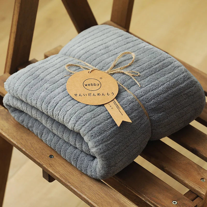 Cette image montre une couverture grise douillette avec une étiquette dorée attachée à l'aide d'un ruban de jute, représentant une marque nommée "Webby".