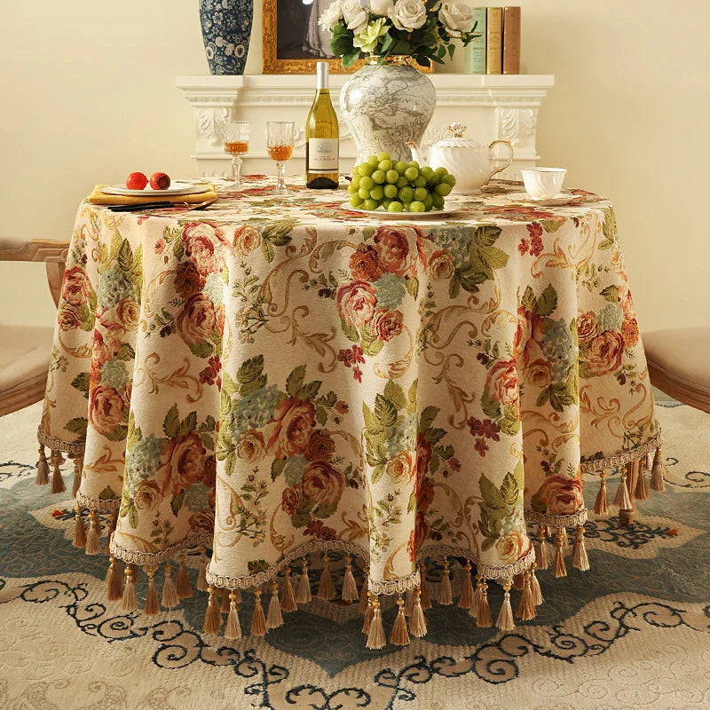Une table drapée d'une nappe fleurie richement ornée de franges et de glands, avec une décoration de vases, de fleurs et de fruits, créant une atmosphère chaleureuse et élégante dans cette pièce.
