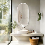 Un aménagement de salle de bain moderne et zen, avec une grande fenêtre offrant une vue sur la nature environnante, un lavabo en vasque blanche posé sur une surface de travail épurée, un miroir ovale lumineux et des accessoires de salle de bain à l'esthétique naturelle.