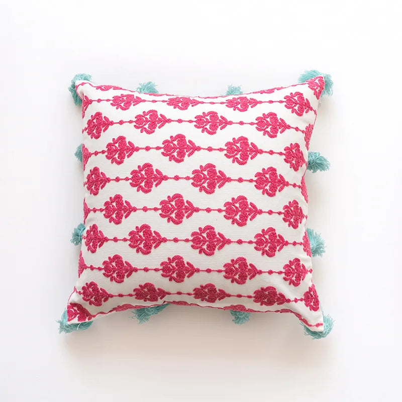 Coussin décoratif blanc et rose foncé avec des motifs floraux et des pompons bleus sur les coins.