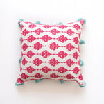 Coussin décoratif blanc et rose foncé avec des motifs floraux et des pompons bleus sur les coins.