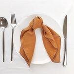 Un joli linge de table orange clair, posé sur une assiette blanche, accompagné d'une fourchette et d'un couteau en acier inoxydable.