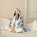 Une jeune femme portant un manteau blanc chaud avec des motifs géométriques noirs et blancs, assise sur un canapé blanc confortablement enveloppée dans une couverture moelleuse.