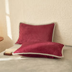 Coussin en velours côtelé de couleur rouge bordeaux posé sur un lit, avec une finition de passepoil grise lui donnant un look élégant et confortable.