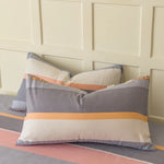 Des oreillers colorés et rayés dans des tons de gris, orange et rose, placés devant un mur de panneaux blancs, créant une ambiance douce et harmonieuse.