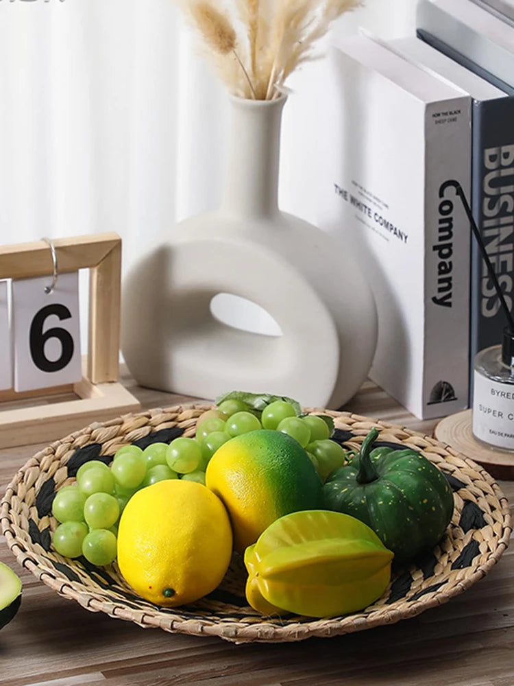 Plateau de fruits et légumes colorés - citrons jaunes, raisins verts, poivrons verts et lime - disposés sur un plateau en osier, avec des livres et un vase blanc en arrière-plan.