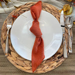 Un arrangement automnal de couverts sur un tapis de fibres végétales, avec une serviette de couleur rouille nouée sur une assiette blanche.