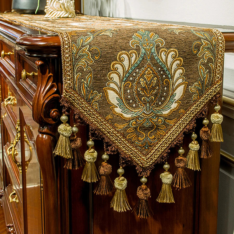 Élégante nappe décorative marron et dorée, ornée de motifs floraux élaborés et de glands frangés, reposant sur une imposante commode en bois.
