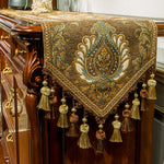 Élégante nappe décorative marron et dorée, ornée de motifs floraux élaborés et de glands frangés, reposant sur une imposante commode en bois.