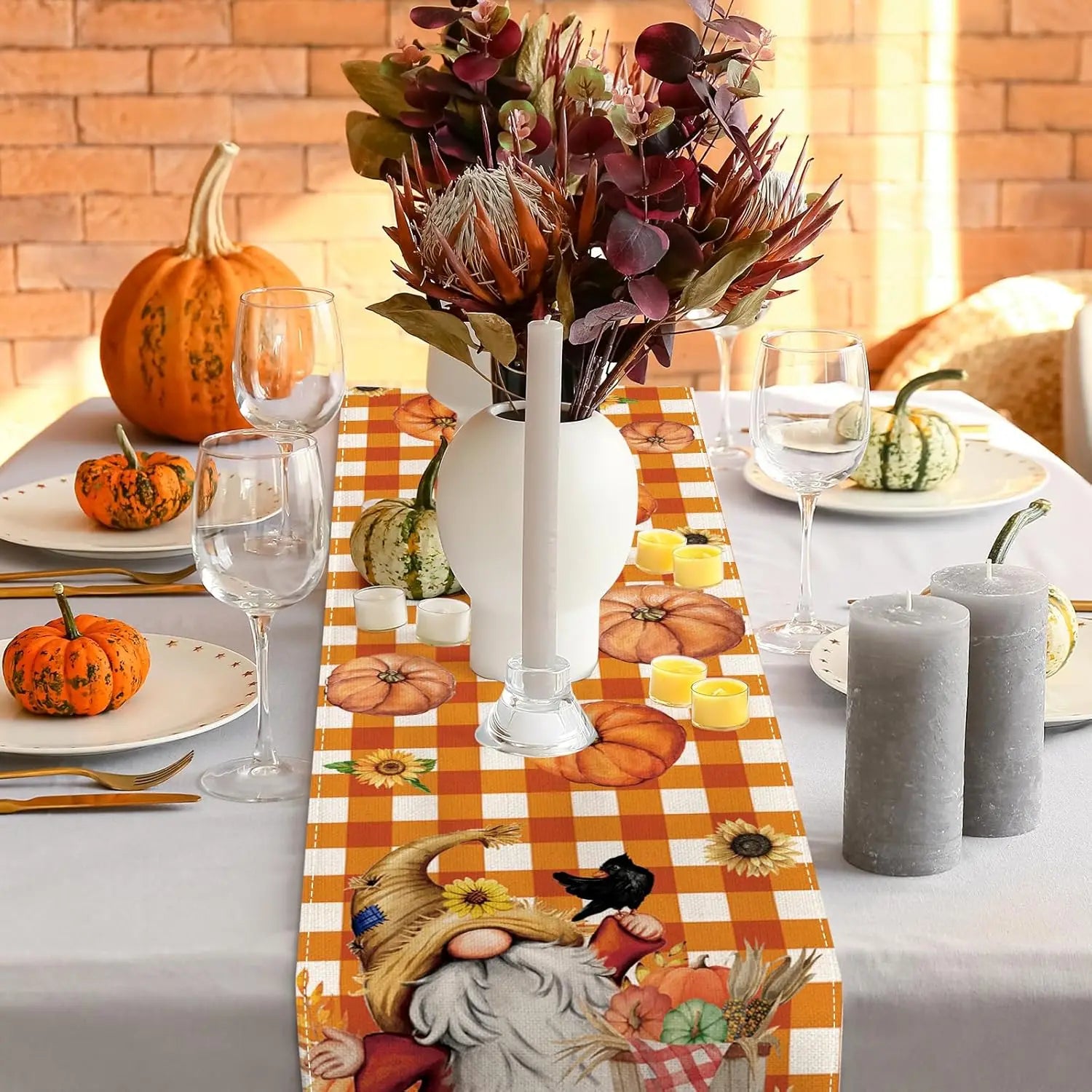 Cette image montre une jolie décoration de table d'automne avec un bouquet de fleurs automnales, des citrouilles, des chandelles et des verres à vin sur un napperon à carreaux oranges et blancs.