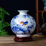 Un vase en porcelaine blanche décoré de motifs floraux bleus et de deux oiseaux colorés, reposant sur un support en bois.