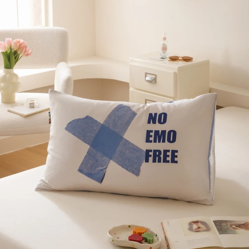 Un coussin décoratif avec le texte "No emo free" imprimé dessus, posé sur une surface blanche à côté d'une fenêtre, de fleurs roses et d'autres éléments de décoration.