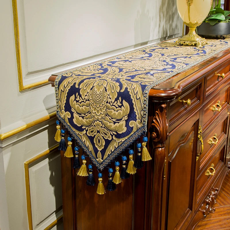 Magnifique chemin de table en jacquard bleu et doré, avec des motifs floraux complexes, des glands et des pompons, reposant sur un élégant meuble en bois d'acajou, créant une ambiance chaleureuse et sophistiquée.