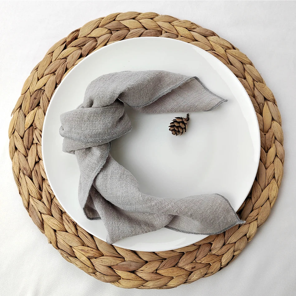 Cercle tressé en fibres naturelles avec une serviette grise enroulée, accompagnée d'une pomme de pin, sur un fond blanc.
