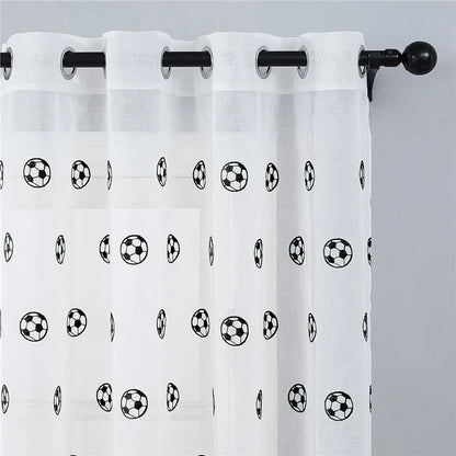 Voilage à motifs football suspendu sur une tringle noire, accroché à une fenêtre sur fond de mur gris.