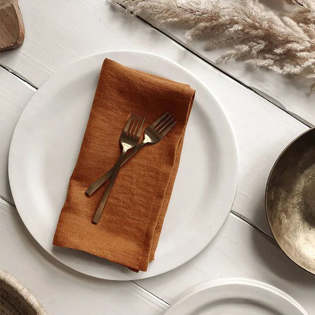 Assiette blanche avec serviette en tissu orange foncé et couverts en métal sur une table en bois, avec des décorations florales.