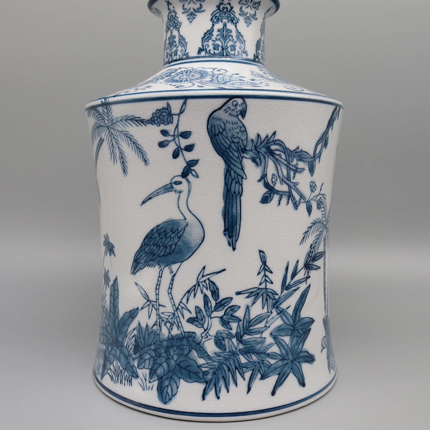 Un vase en porcelaine orné de motifs bleus et blancs représentant des oiseaux, des feuilles de palmier et une végétation luxuriante, créant une scène exotique et sophistiquée.