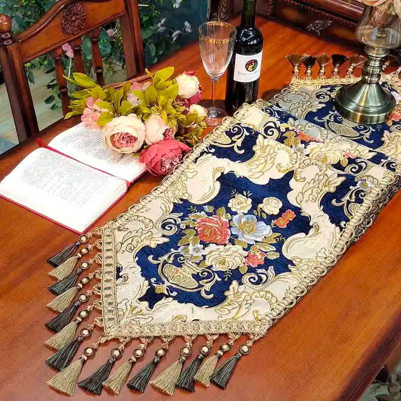 Élégante nappe en satin brodé bleu et crème, accompagnée de fleurs luxuriantes, d'une bouteille de vin et d'un livre ouvert, créant une scène chaleureuse et raffinée pour une soirée conviviale.