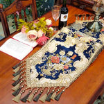 Élégante nappe en satin brodé bleu et crème, accompagnée de fleurs luxuriantes, d'une bouteille de vin et d'un livre ouvert, créant une scène chaleureuse et raffinée pour une soirée conviviale.