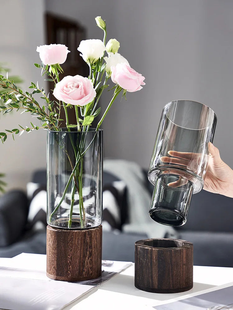 Un arrangement floral élégant et épuré, composé de roses roses et de fleurs blanches délicates, mis en valeur par un vase en verre sur une base en bois brut.