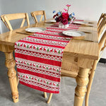 Cette image présente une table en bois avec des chaises et un chemin de table décoré de motifs de rennes rouges et de flocons de neige, avec un bouquet de fleurs rouges sur le dessus.