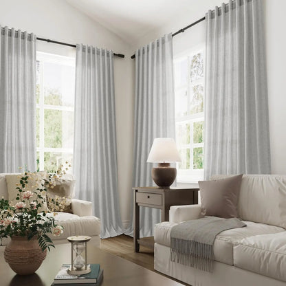 Vue d’ensemble d’un salon moderne et cosy avec de grands voilages gris clair suspendus aux fenêtres, baignés de lumière naturelle.