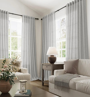Vue d’ensemble d’un salon moderne et cosy avec de grands voilages gris clair suspendus aux fenêtres, baignés de lumière naturelle.