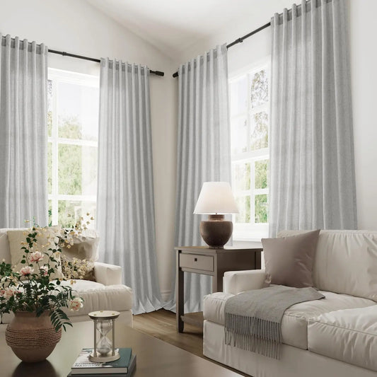 Vue d’ensemble d’un salon moderne et cosy avec de grands voilages gris clair suspendus aux fenêtres, baignés de lumière naturelle.