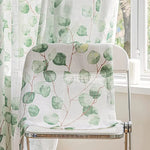 Voilage feuillage vert tombant élégamment sur une chaise blanche placée devant une fenêtre.