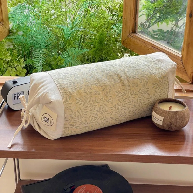 Un sac de voyage en tissu délicat avec un motif de feuilles, posé sur une table en bois avec une bougie allumée et un lecteur de vinyles à proximité, le tout entouré d'une végétation luxuriante visible par la fenêtre.