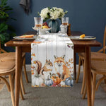 Une nappe de table en tissu avec des motifs de renards, d'écureuils et de citrouilles, entourée de verres à pied et d'un bouquet de fleurs, sur une table en bois dans un environnement douillet et automnal.