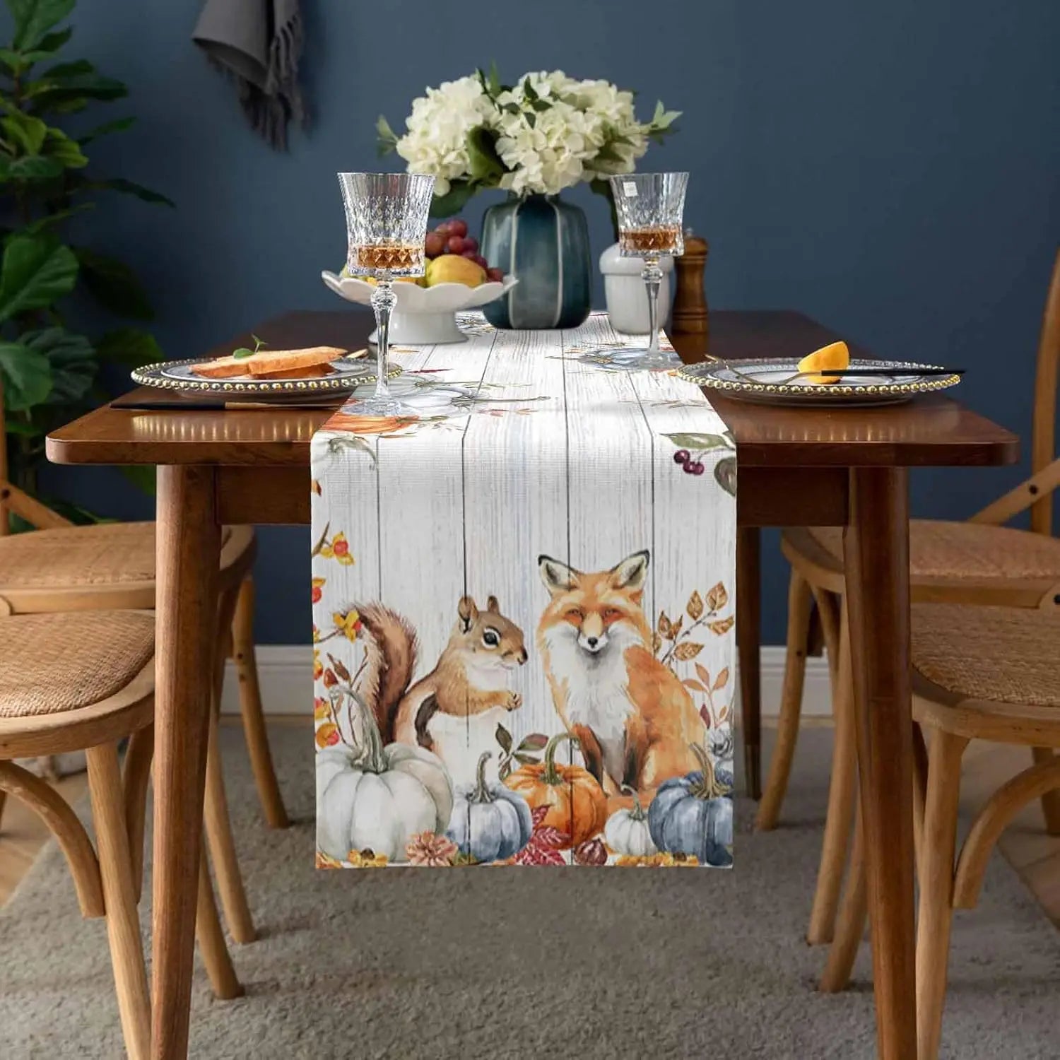Une nappe de table en tissu avec des motifs de renards, d'écureuils et de citrouilles, entourée de verres à pied et d'un bouquet de fleurs, sur une table en bois dans un environnement douillet et automnal.