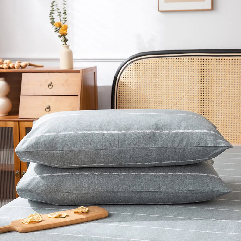 Deux coussins gris en velours, posés sur un lit avec une table de chevet en bois et un fauteuil en rotin en arrière-plan.