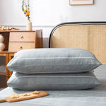 Deux coussins gris en velours, posés sur un lit avec une table de chevet en bois et un fauteuil en rotin en arrière-plan.