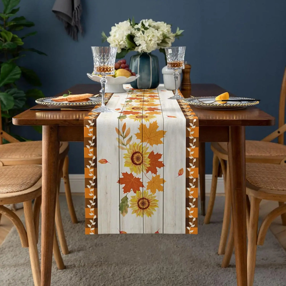 La table de salle à manger a un joli décor automnal avec un chemin de table imprimé de feuilles d'érable jaunes et orange, des fleurs blanches dans un vase et des verres à vin.