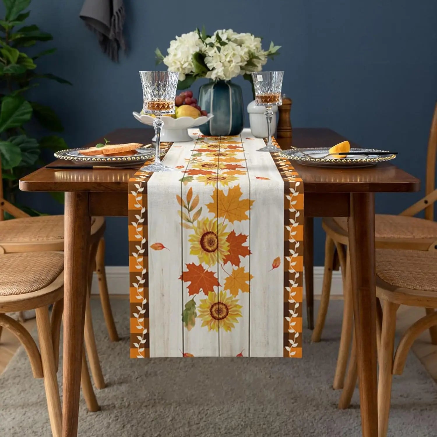 La table de salle à manger a un joli décor automnal avec un chemin de table imprimé de feuilles d'érable jaunes et orange, des fleurs blanches dans un vase et des verres à vin.