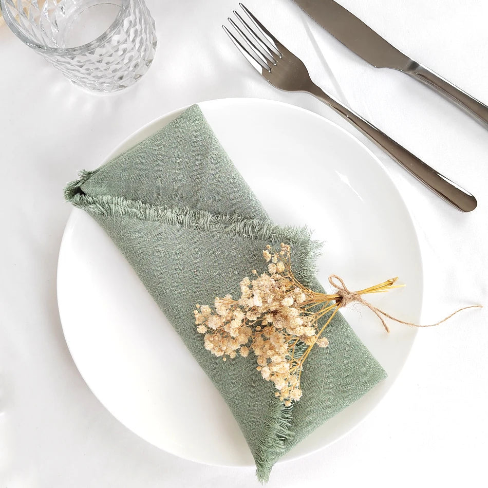 Une serviette de table en lin vert ornée de fleurs séchées, posée sur une assiette blanche avec des couverts en métal.
