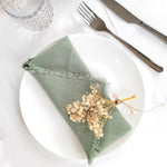 Une serviette de table en lin vert ornée de fleurs séchées, posée sur une assiette blanche avec des couverts en métal.