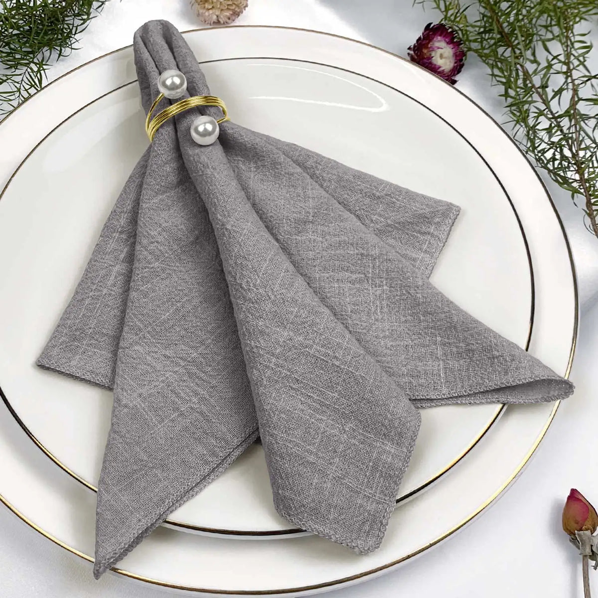 Serviette grise avec anneaux dorés, posée sur une assiette blanche, entourée de fleurs et de feuillage décoratifs.