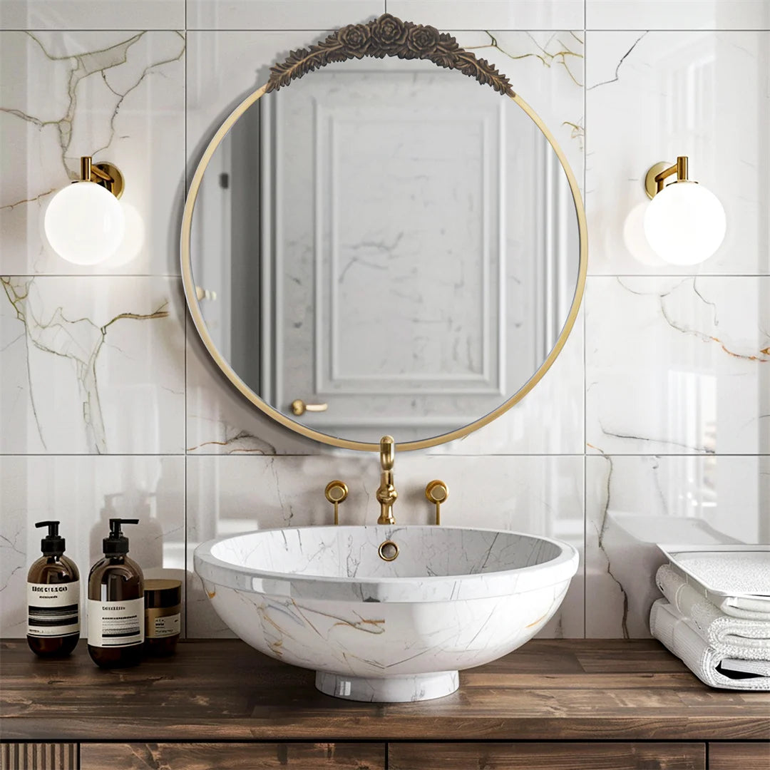 Miroir rond doré avec appliques dorées éclairant un lavabo en marbre blanc posé sur un meuble en bois sombre dans une salle de bain élégante aux carreaux de marbre.