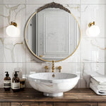 Miroir rond doré avec appliques dorées éclairant un lavabo en marbre blanc posé sur un meuble en bois sombre dans une salle de bain élégante aux carreaux de marbre.