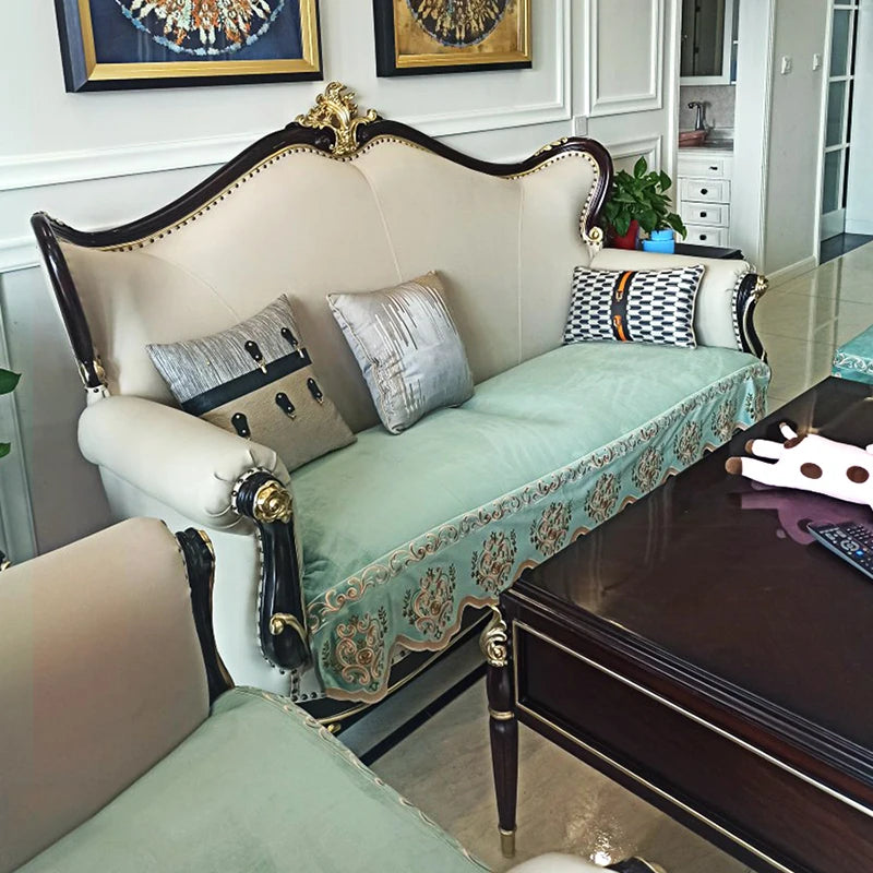 Un élégant canapé de style baroque, avec des détails ornementaux en bois foncé et un rembourrage vert pastel, décoré de coussins aux motifs modernes et géométriques, le tout placé dans un intérieur avec des murs blancs et des éléments de décoration vintage.