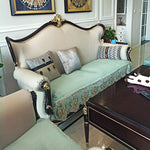 Un élégant canapé de style baroque, avec des détails ornementaux en bois foncé et un rembourrage vert pastel, décoré de coussins aux motifs modernes et géométriques, le tout placé dans un intérieur avec des murs blancs et des éléments de décoration vintage.