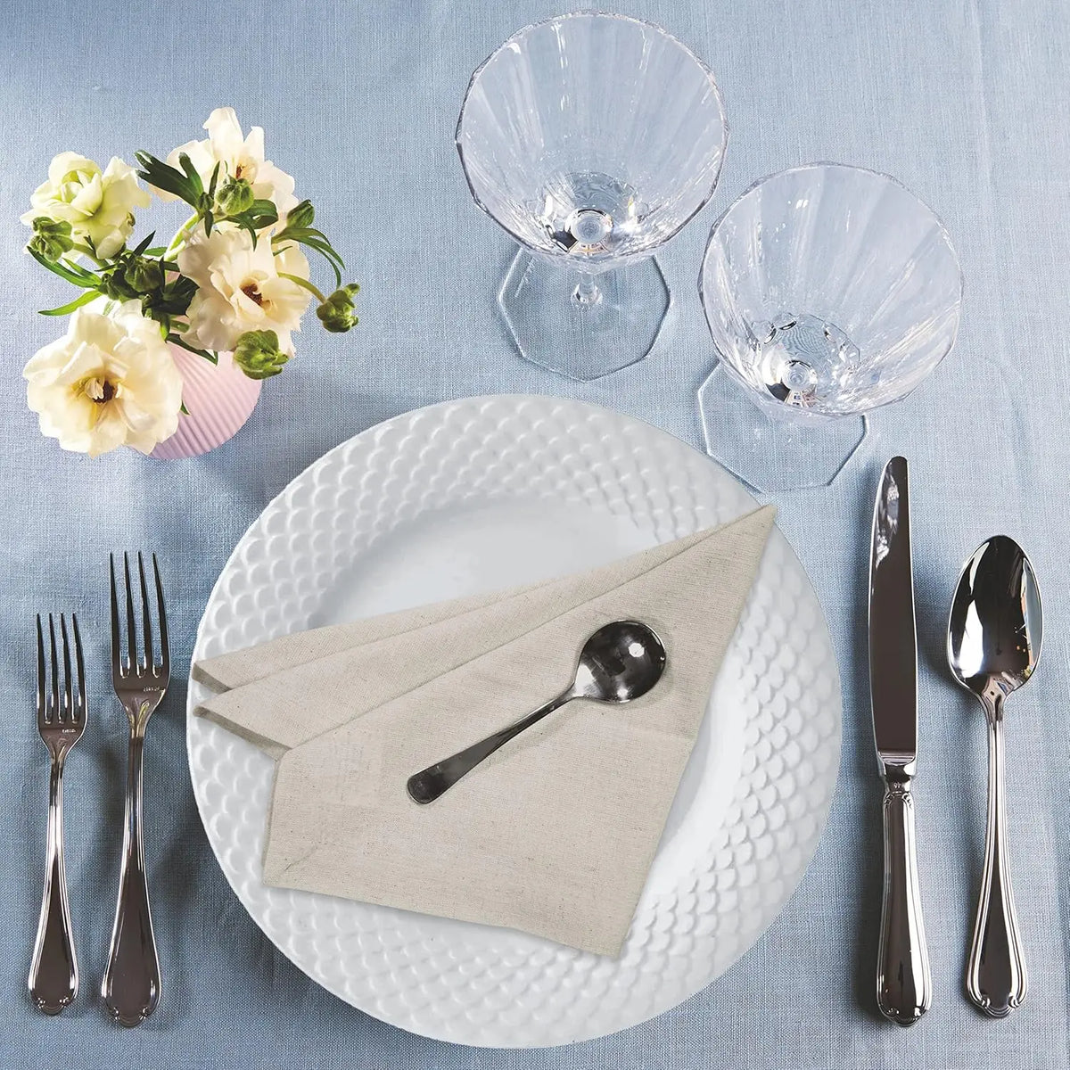 Une élégante table de repas avec une assiette blanche texturée, des couverts argentés, des verres de cristal et un arrangement floral.